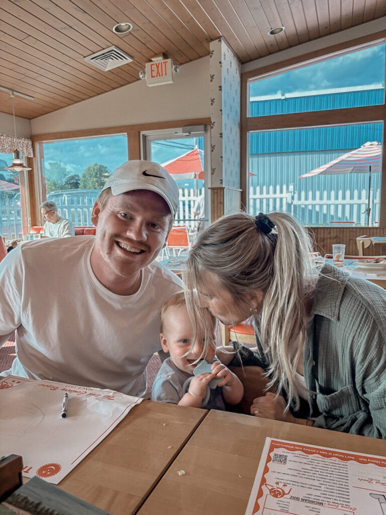 Husband and wife sitting at a table, husband smiling at camera, wife kissing their baby