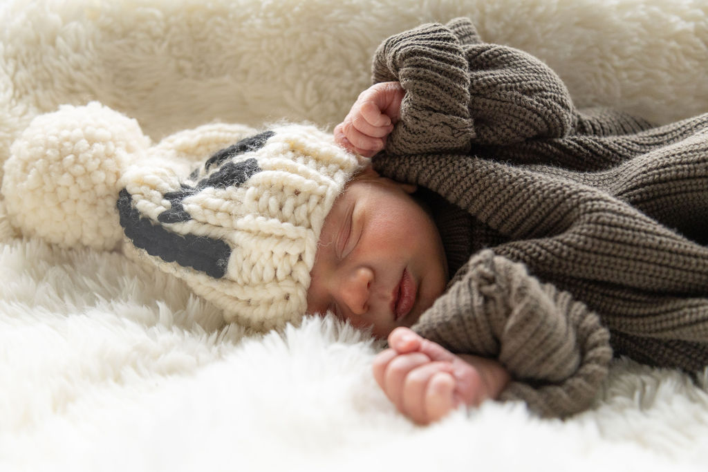 Newborn sleeping wearing a white knit hat and oversized green sweater