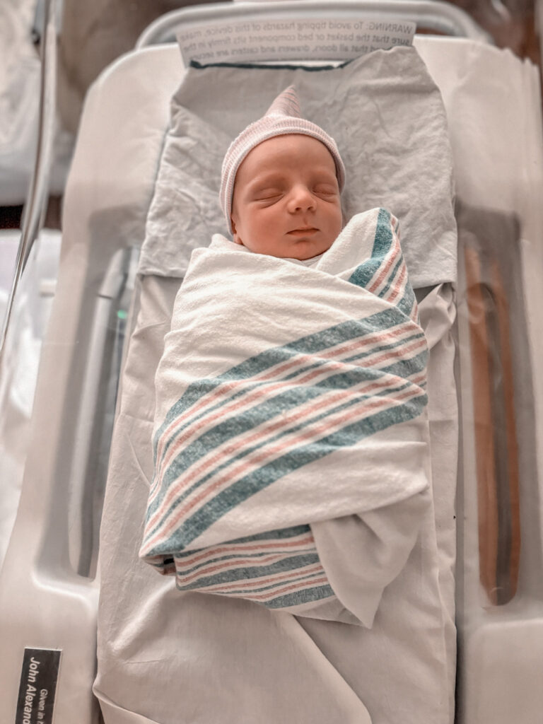 newborn baby sleeping wrapped in a white swaddle with green and pink stripes wearing a white knit hat.