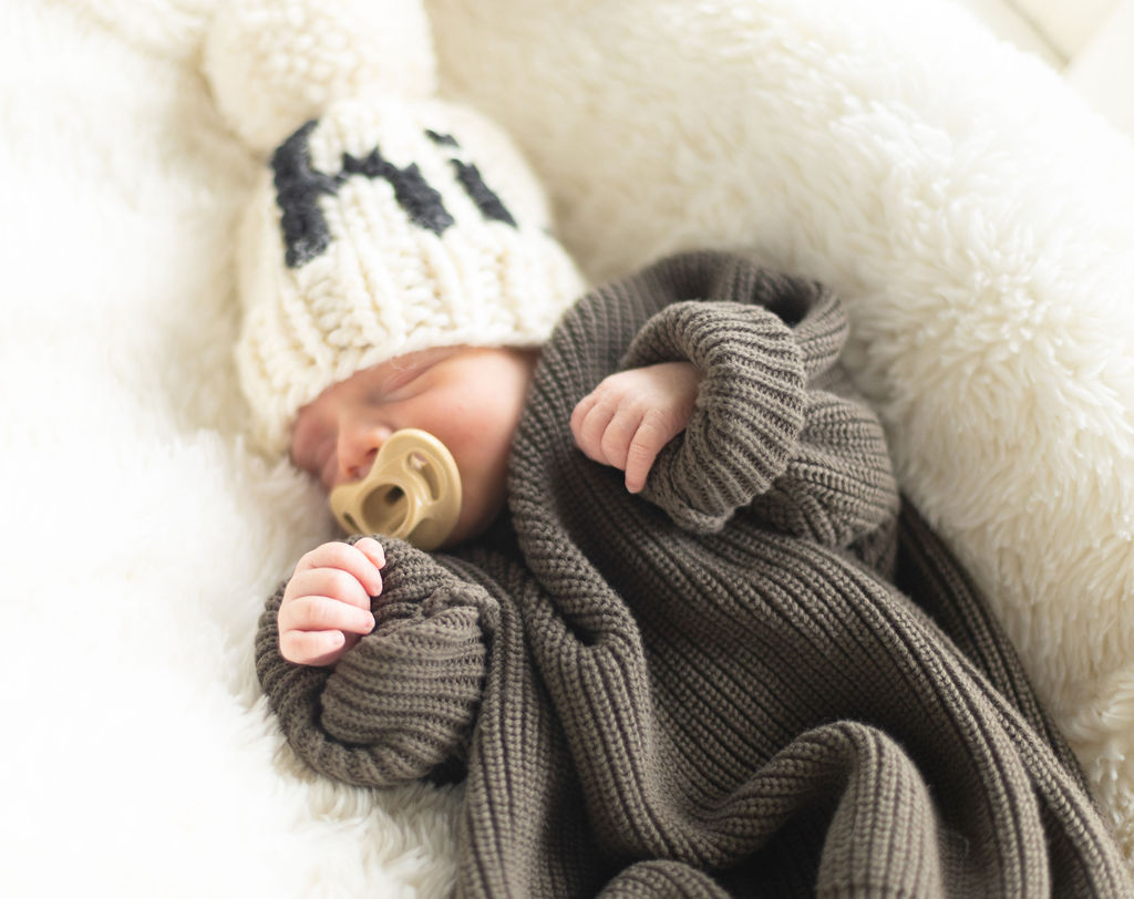 sleeping newborn dressed in a green oversized sweater, white knit hat and a tan pacifier.