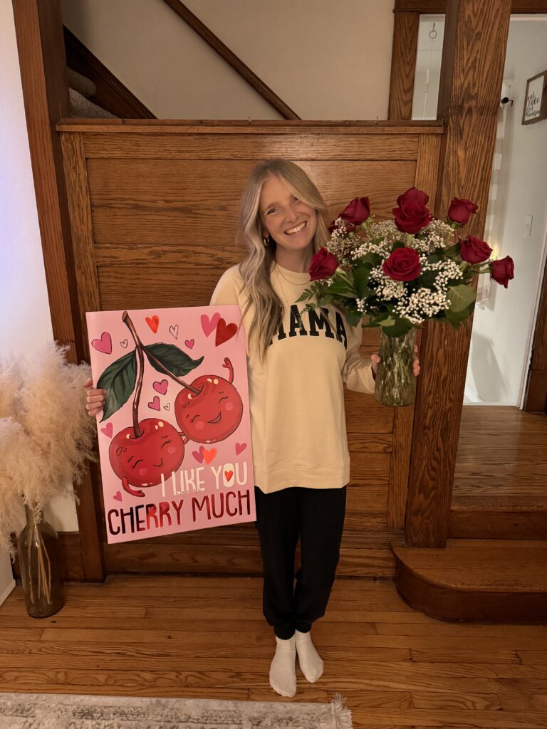 Mama smiling wearing a mama sweatshirt carrying a giant card and roses