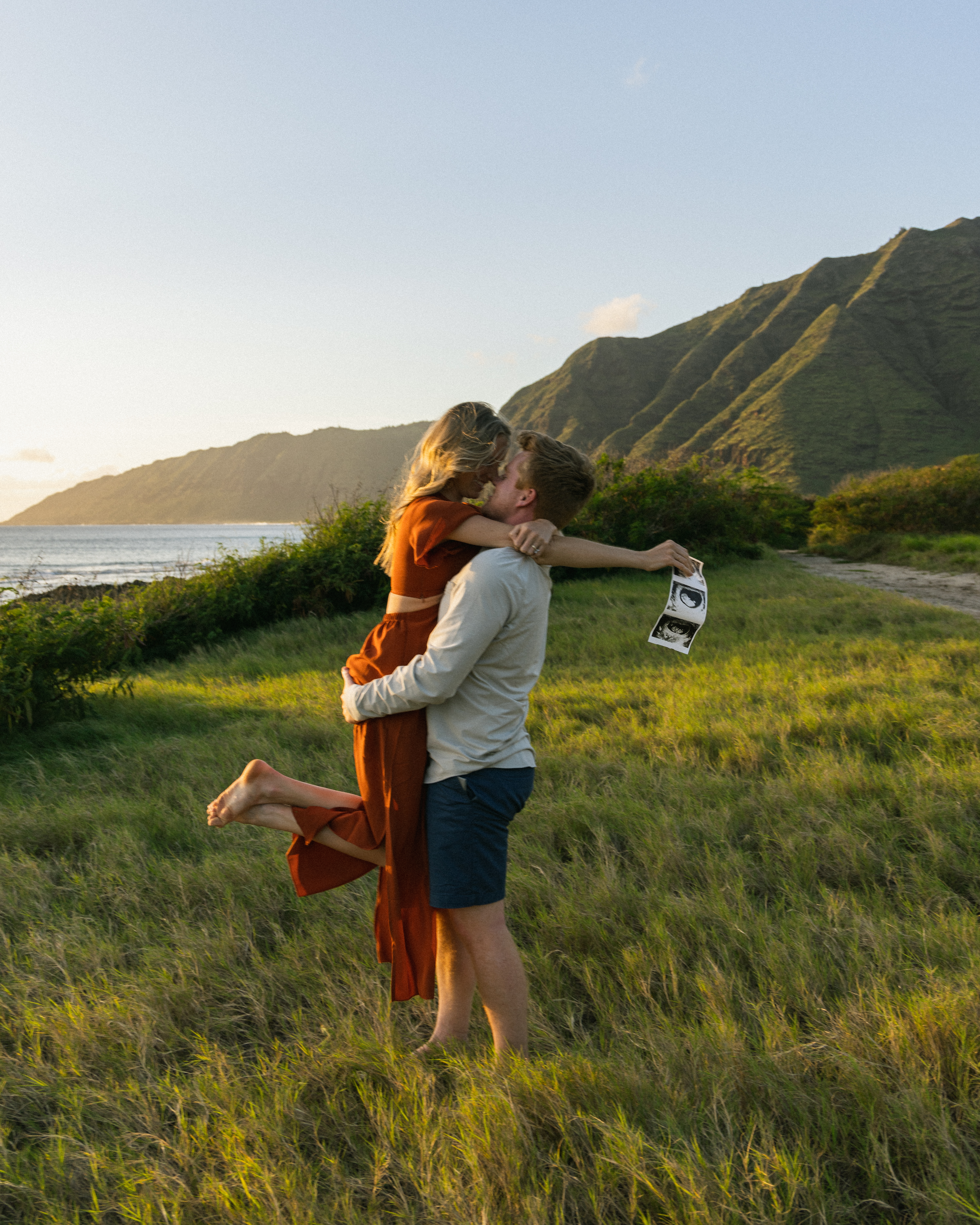 Husband picking wife up in a beautiful green field, she is holding ultrasound pictures