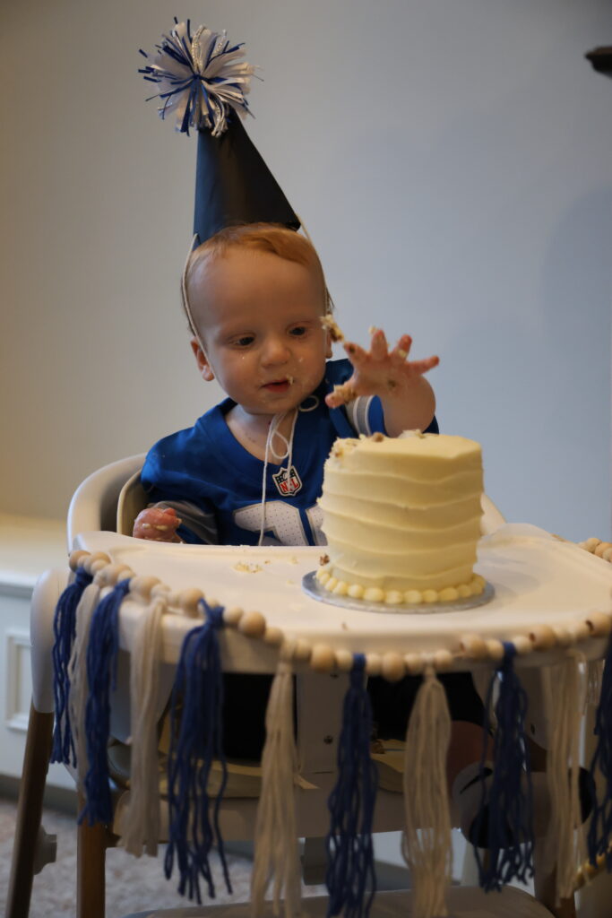 1 year old boy with party hat on reaching for a bite of his smash cake