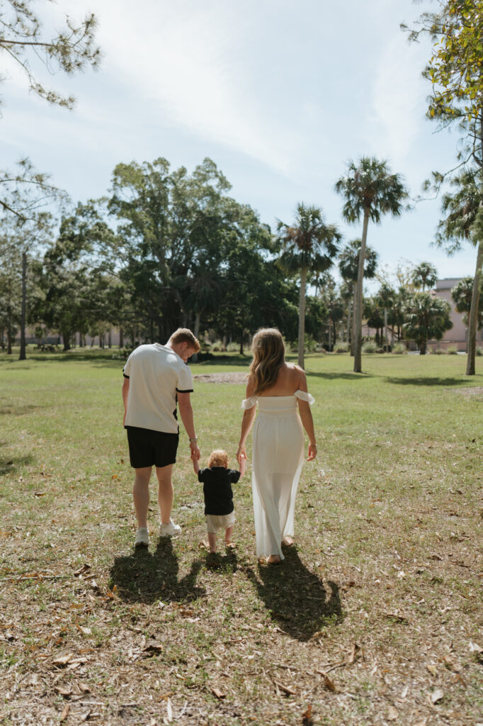 Mom and dad holding their one year old son’s hands walking away from the camera