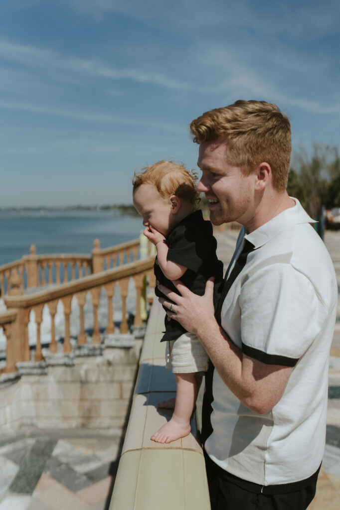 Profile view of a dad holding his one year old son smiling looking into the ocean 
